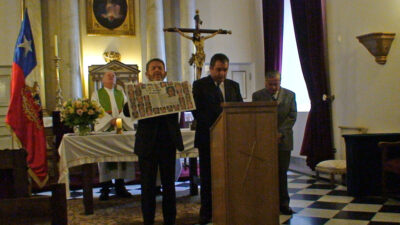 priest stands behind altar table while men pray at lectern holding newspaper pictures of the 33 Chilean miners