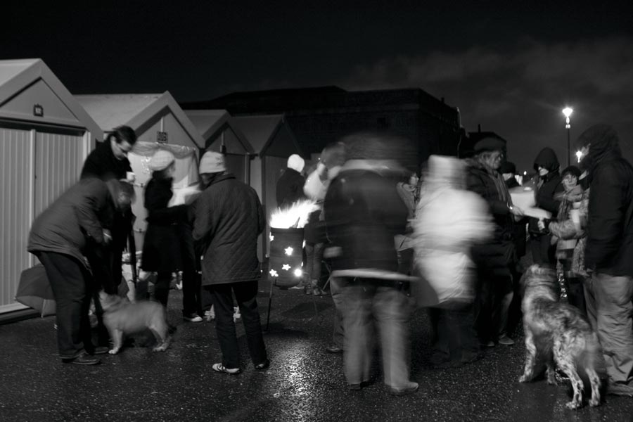 People gather outside beach huts at night round a brazier
