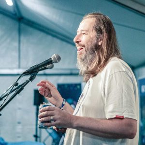 Smiling man with long hair and beard speaking into microphone in festival tent