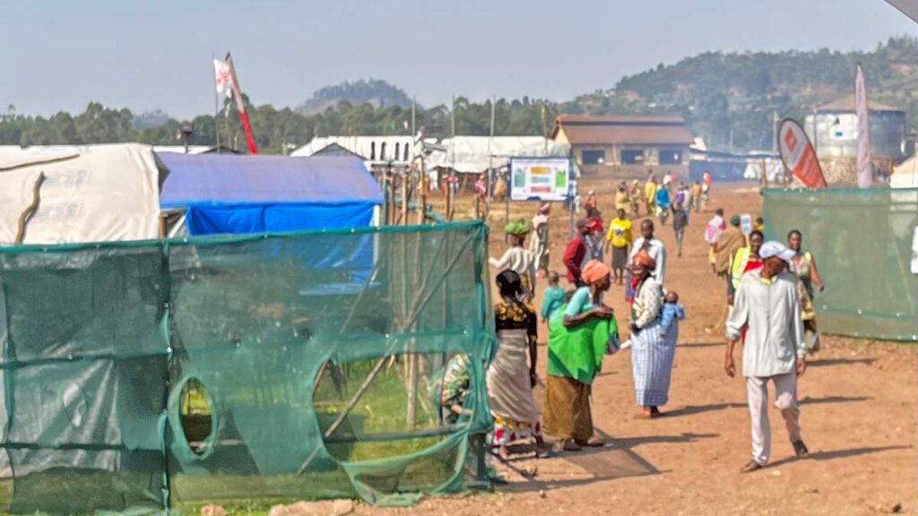 People mill around the fence of a refugee camp on dusty red road