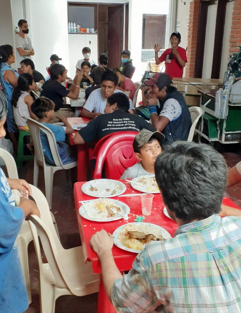 Twenty or so people of varying ages seated around plastic tables with food, listening to a Bolivian man speak