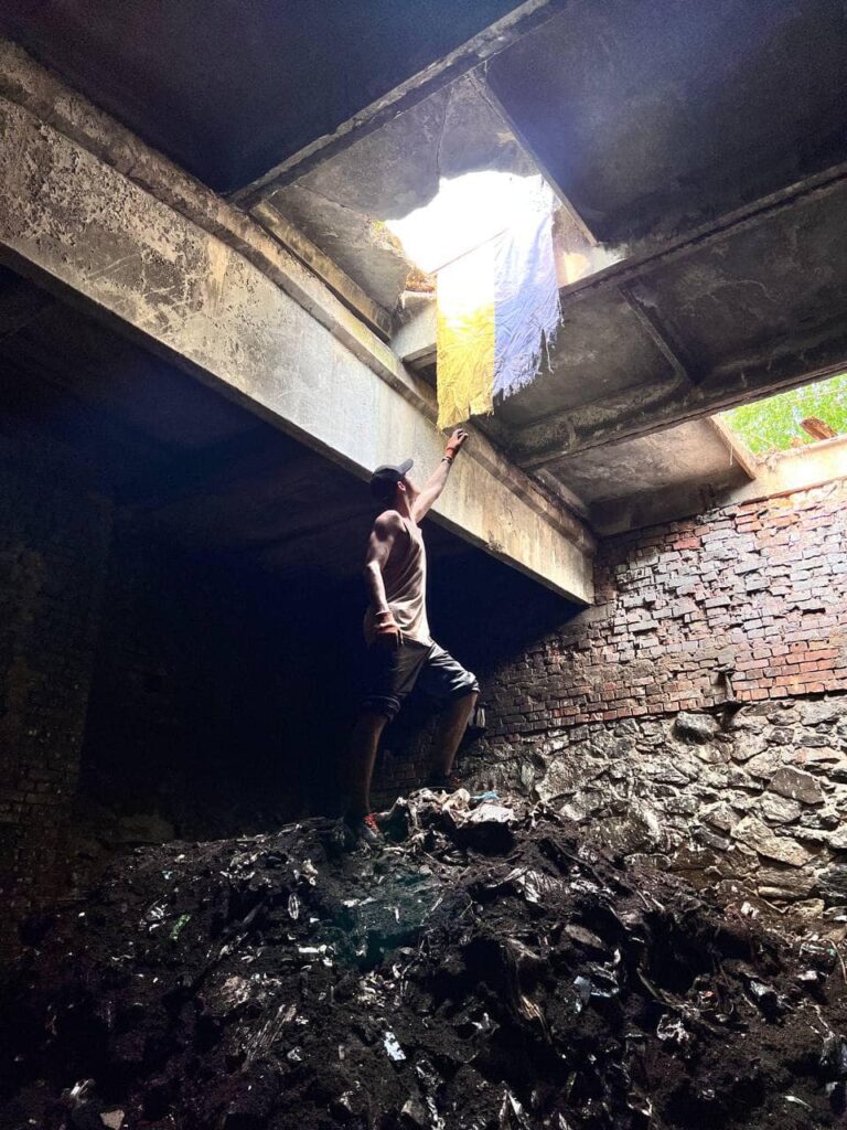 young man stands on pile of rubble in basement, reaching up for tattered Ukrainian flag hanging through a hole in the ceiling