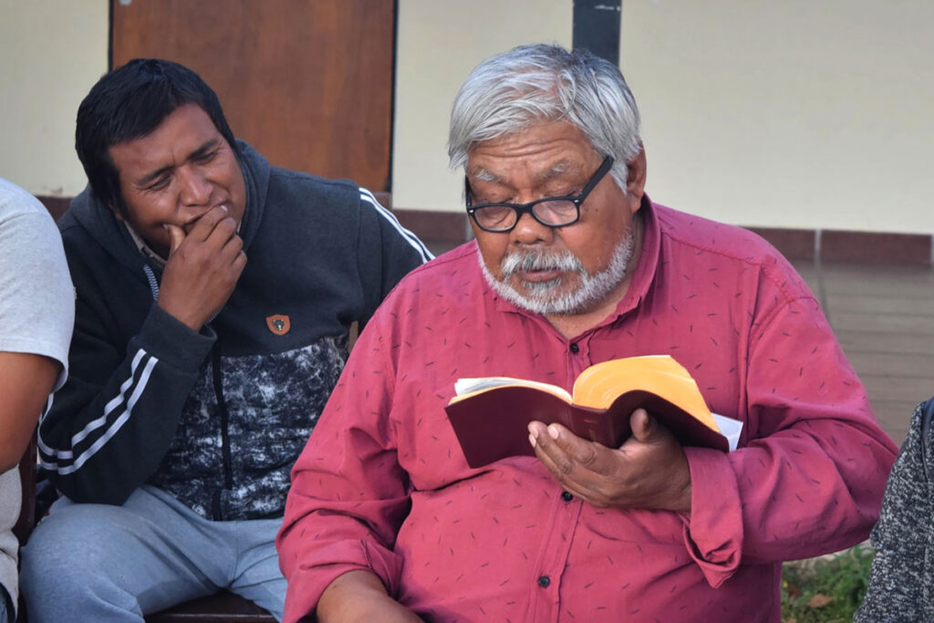 indigenous elder with white beard reads from Bible while others listen