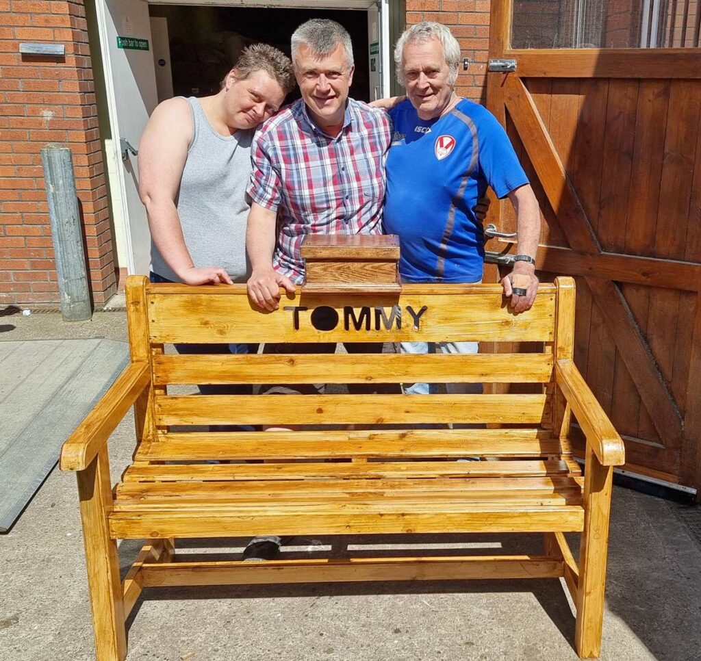Garry and shed members stand behind a garden bench with cut-out lettering saying "TOMMY"
