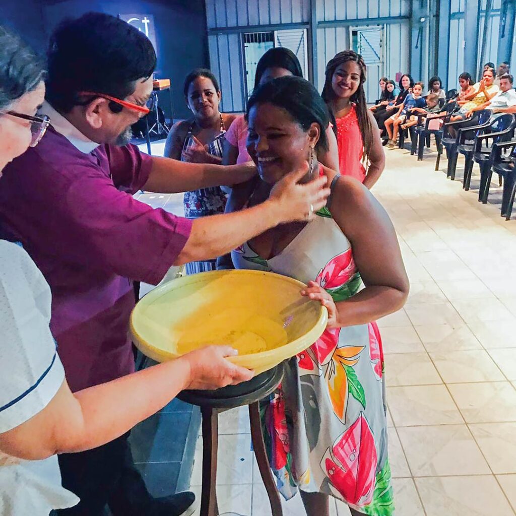 Bishop in purple clerical shirt splashes water over a smiling Brazilian woman