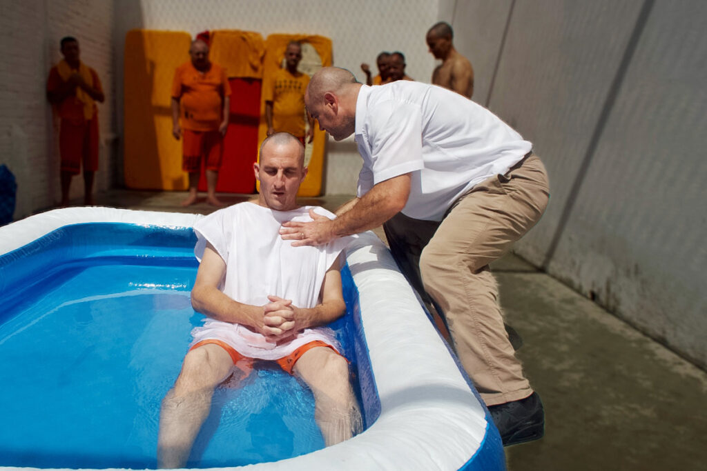 man in short sleeved shirt holds another man in white and orange, sitting in an inflatable pool