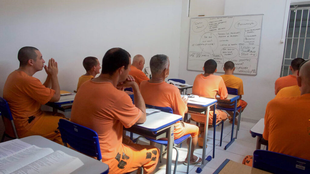 a dozen men in orange t-shirts and trousers sit in a sparse classroom studying the whiteboard