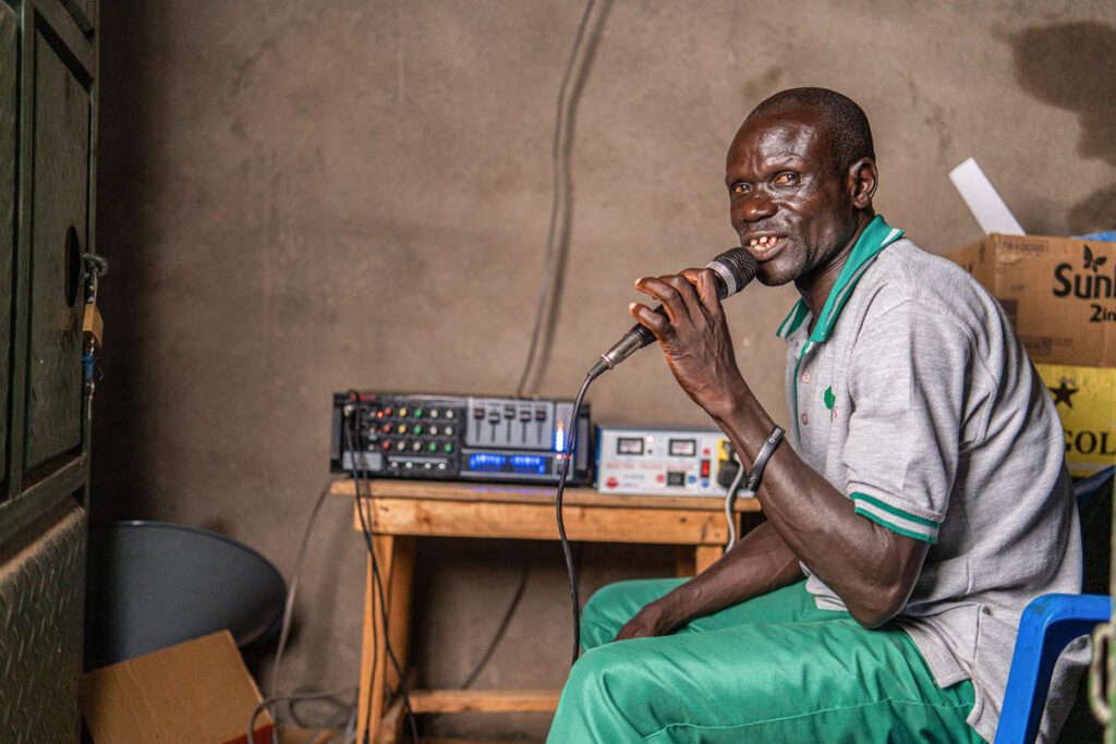 South Sudanese trauma healing worker speaking into microphone plugged into sound mixer atop a small wooden table against a mud wall