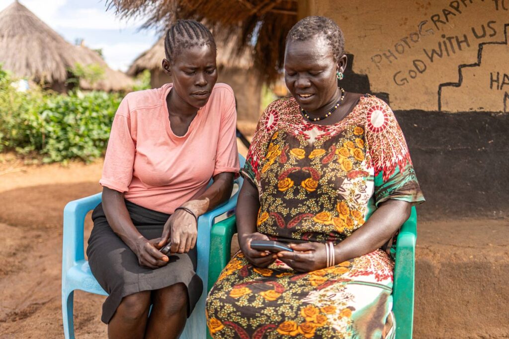 two South Sudanese women sit outside a thatched hut listening to an audio player