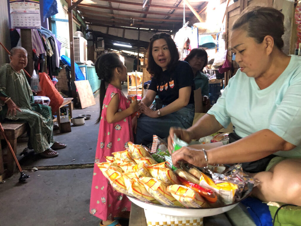 Thai woman talks to a young girl and another Thai woman sorting packaged foodstuffs on a tray