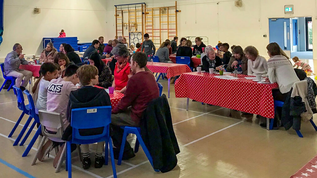 people of all ages seated in school hall around tables with bright red and white polkadotted tablecloths