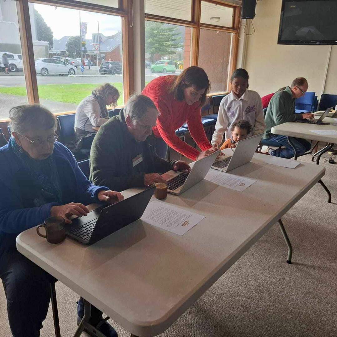 people use rows of laptops on large tables in community centre-type room
