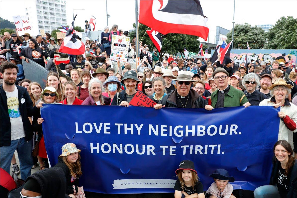 crowd gathers behind banner reading "LOVE THY NEIGHBOUR HONOUR TE TIRITI"