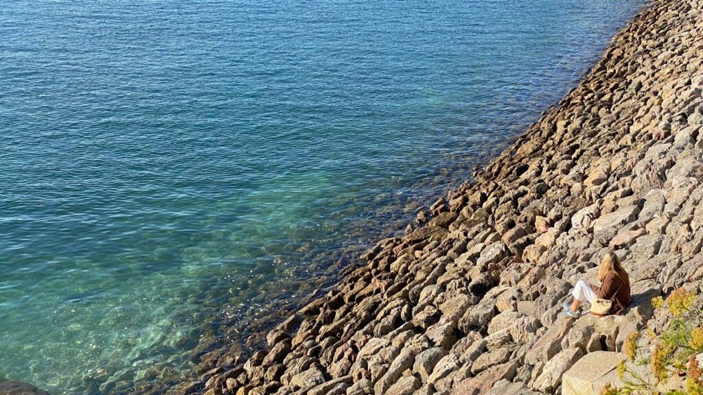lone woman sitting on rocky shore with blue-green sea