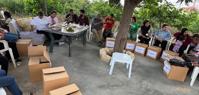 Lebanese folk talk together, sit in a circle of chairs with large cardboard boxes in the centre
